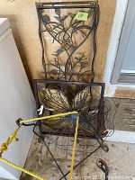 Two black metal garden wall art pieces: tall vine-leaf design and square gold flower design leaning against wall and cooler.