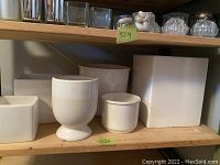 Front view showing six assorted white ceramic planters on a wooden shelf. Various shapes include round, square, and pedestal styles.