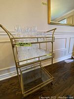 Front view of brass finish metal bar cart displaying top two shelves filled with cut crystal glassware