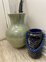 Green ceramic vase with wide body and flared top, with sticker indicating 19 inches tall and price $140, positioned beside black ceramic lantern with blue bead handle.