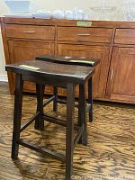 Set of two dark brown wood stools with rectangular seats and reinforced angled legs on hardwood floor in front of wooden cabinet.