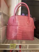 Front view of pink croc-embossed leather handbag with two rounded handles, displayed upright on a shelf.