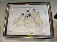 Framed artwork showing Inuit figures seated and looking upwards, framed in a silver metallic frame.