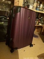 Full front and side view of the hard case luggage standing upright on a basement floor, showing maroon color, vertical ridges, and wheels.