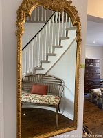 Full view of mirror leaning against wall, showing carved gold frame and reflection of staircase and furniture.