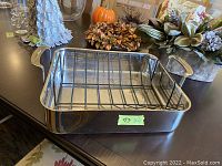 Angle view showing large stainless steel roasting pan with handles and black roasting rack inside on dark surface with decorative items in background.