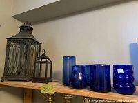 Photo of a wooden shelf holding two metal lanterns and five blue glass vases of various shapes and sizes arranged side by side.