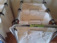 Close image showing three wheeled laundry bins with beige fabric removable bags on metal frames arranged in a row.