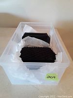 Set of two black clutches placed inside a clear plastic storage container with white tissue paper cushioning them. One clutch has a studded surface and the other has a smooth surface, both rectangular in shape.