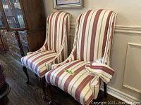 Two custom wingback accent chairs showing striped upholstery and extra fabric material on one armrest, dark wooden legs, placed on a wood floor.
