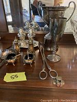 Photo showing silver trophy, goblet, condiment holders with spoons, salt and pepper shakers on tray, sugar tongs, and lighters