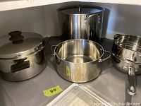 Four stainless steel pots displayed on a shelf. Includes large stockpot, double boiler base pot, large pot with side handles, and smaller pot with long handle. Missing lids on some items.
