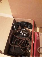 Black rotary dial telephone with coiled cord inside an opened cardboard box on the floor.