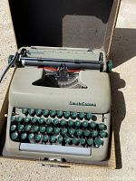 Vintage gray Smith Corona typewriter with teal keys inside the vintage open carrying case on concrete surface.