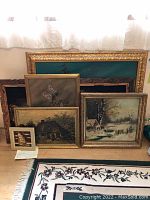 Photo of multiple framed paintings leaning against a wall, including ship and landscape artworks plus handwritten note