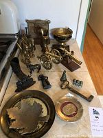 Overall view of the brass and lead decorative items arranged on a table showing cannon, flintlock pistol, horses, bowls, boot vase, bookend and assorted small pieces.