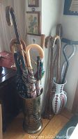 Photo showing large brass embossed cylindrical umbrella stand filled with umbrellas with various curved handles, placed next to a Ukrainian decorative vase holding wooden canes.