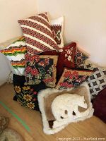 Photo shows multiple cushions stacked against a wall, featuring needlepoint floral and geometric designs plus fur polar bear cushion
