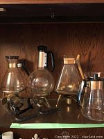 Four different Mid-Century Modern glass coffee carafes displayed on a shelf with three metal stands underneath.