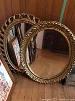 Two round mirrors leaning against a wall with detailed gold-colored carved frames, one with shell/leaf edge pattern and the other with open scroll design.