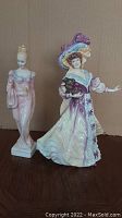 Two Royal Doulton porcelain figures: Daphne in pink dress and Lily in an elegant cream and lavender dress holding a dog.