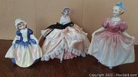 Three figurines shown: two glossy porcelain Royal Doulton figurines and one fabric skirt doll with porcelain parts.