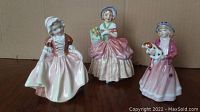 Three Royal Doulton porcelain figurines on a wooden surface with a plain cardboard background, showing front view of each figure.