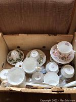 Box containing multiple teacups and saucers viewed from above showing various bottom stamps including Royal Albert and Queens Staffordshire.