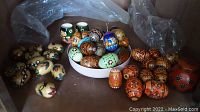 Photo showing approximately 30 wood Ukrainian decorated eggs with varied floral and folk art patterns arranged in a cardboard box along with two painted wooden cup figures and a round wooden container.