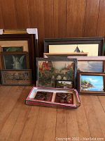 Several framed vintage paintings and wall plaques arranged against a wooden paneled wall on the floor, showing a variety of artworks including angel, village scenes, and a boxed set of relief plaques depicting a motorcycle and sidecar.