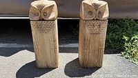 Two hand carved wooden owl statues with square columnar bodies and carved feather detailing, standing on pavement outside.