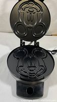 Open waffle maker showing nonstick cooking plates with Mickey Mouse face design on top and bottom plates, black finish, power cord visible.