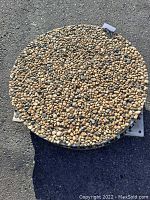 Top view of one concrete garden aggregate round showing tan, brown, black, and gray smooth stones in concrete matrix on asphalt background.