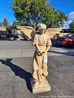 Front view of medium weight resin composition garden angel statue with wings spread, standing on rectangular base, outdoors with trees and cars in background.