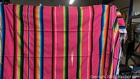 Photo showing the full vibrant Mexican serape blanket held vertically, highlighting multicolored horizontal stripes and the fringed bottom edge.