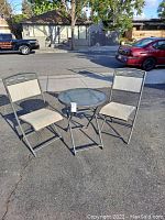 Photo of outdoor metal bistro table with glass top and two matching folding chairs with beige outdoor fabric upholstery set on asphalt outdoors
