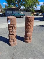 Photo of two large red-brown resin tiki god totem poles placed outdoor on asphalt with street and cars background.
