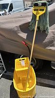Yellow Rubbermaid Wave Break mop bucket with wringer and wooden-handled mop with yellow mop head attachment and worn mop head, set outdoors.