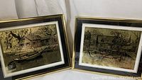 Pair of framed foil-etched artworks side-by-side showing gold/black etchings of waterfront cottages and boats