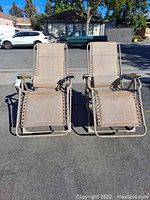 Front view of two tan zero gravity lounge chairs side by side on asphalt surface outdoors.