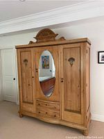 Full front view of antique wood armoire showing three doors and oval mirror on center door, along with decorative carved top.
