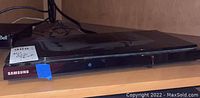 Front view of Samsung BluRay player on wooden shelf showing brand and power button with blue tape on corner.