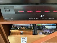 Front panel view of the black R1 CAV 6 Multi-zone controller showing six red LED indicators and power switch on the left, placed above a shelf with various DVDs or CDs.