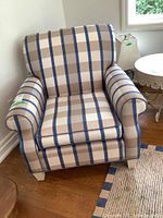 Frontal view of upholstered occasional chair with plaid fabric and fabric arm protectors on rolled armrests. Positioned near window and round side table.