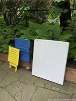 White plastic folding table and yellow folding table folded, blue fabric folding chair folded, leaning against planter box outdoors.