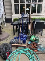 Full view showing assembled gardening tools including shovels, rakes, watering cans, hoses and cords placed on patio.