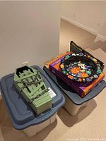 Two closed plastic storage bins containing Halloween decorations, one with a green coffin prop on top and a boxed Halloween plate set on the other.