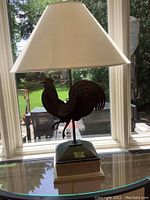 Full view of the metal rooster design table lamp with white lampshade on glass table in front of window, showing overall shape and proportions.