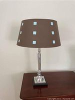 Photo of entire modern table lamp showing brown fabric shade with blue square accents, silver turned metal base, and black square platform base, placed on wooden surface.