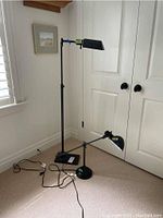 Both lamps positioned on carpeted floor near closed white doors and window with blinds. Floor lamp is taller with rectangular shade; desk lamp is smaller with round shade.